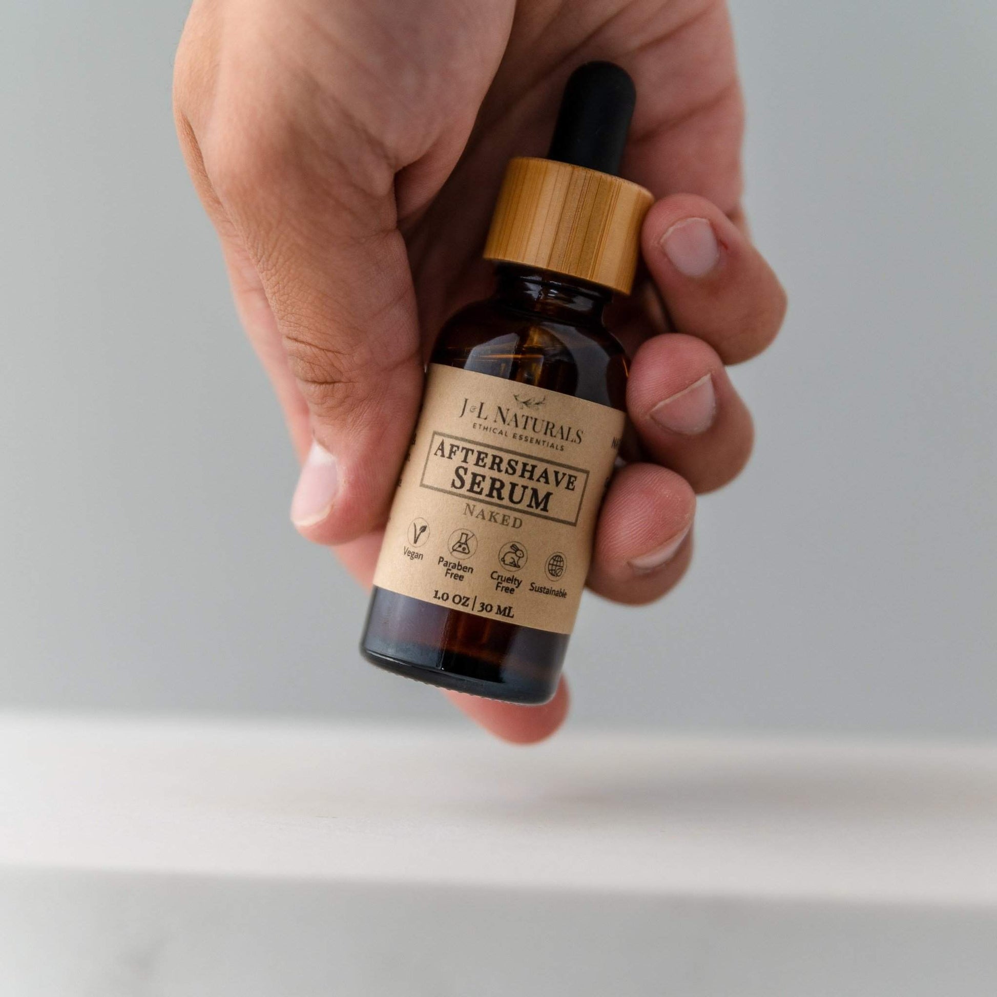 Hand holding a bottle of T.L. Naturals Aftershave Serum with a wooden cap on a light gray background.